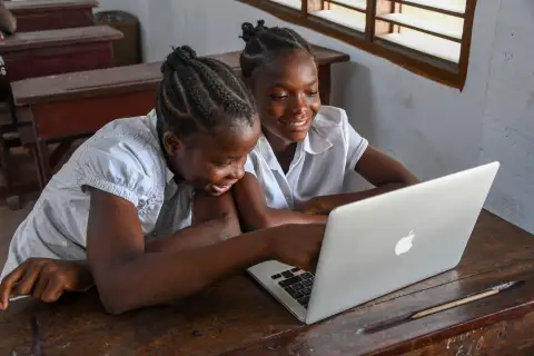 Deux élèves rient devant un ordinateur portable, dans une salle de classe.